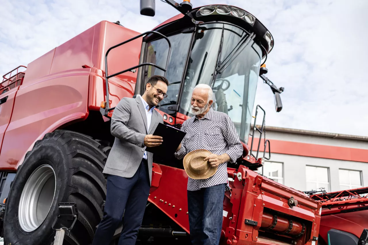 Deux hommes souriants, un en costume et l'autre en chemise, près d'un gros tracteur rouge.