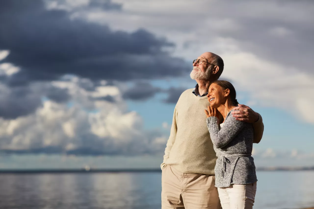 Couple âgé heureux se câlinant au bord de l'eau, nuages en arrière-plan.