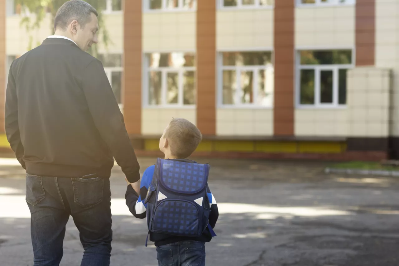 Dos d'un homme et jeune enfant avec sac à dos bleu marchant vers une école.